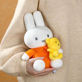 White plush toy with orange outfit holding a yellow plush toy, held by a person wearing a beige sweater.