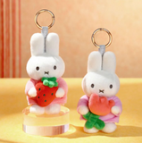 Two plush keychains of a white rabbit holding a strawberry and a flower on a yellow background.