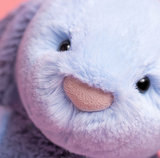 Close-up of a plush stuffed animal face with soft lavender-blue fur, a pink nose, and dark eyes against a pink background.