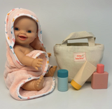 Baby doll wrapped in a hooded towel with baby care products and a bag on a light gray background