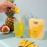 Hand pouring a yellow drink from a pineapple into a glass with a straw, surrounded by a pineapple, passion fruit, and 'mini + me' packaging on a light blue background.