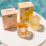 Two glasses of yellow liquid with ice cubes on a white surface, next to 'mini + me' product boxes against a blue water background.