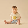 Baby sitting on a beige surface wearing a light-colored shirt with a lemon design and denim shorts.