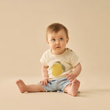 Baby sitting on a beige surface wearing a light-colored shirt with a lemon design and denim shorts.
