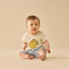 Baby wearing a white shirt with a yellow design and denim shorts on a beige background