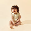 Baby sitting on a beige background wearing a lace romper.