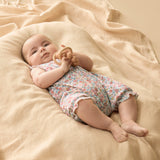 Baby in a floral outfit lying on a beige blanket with a wooden toy.