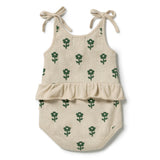 A baby romper in a light beige linen-like fabric with a green floral pattern, featuring tie-shoulder straps and a ruffled waistband, shown on a plain white background.