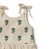 Close-up of a sleeveless baby dress in warm beige linen-like fabric with a repeating green knit flower pattern and tie shoulder straps, shown on a white background.