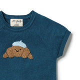 Close-up of a short-sleeve knit top in deep teal blue, featuring a croissant motif with a light blue beret on the chest. The brand label inside the collar reads 'woven + friendly'.
