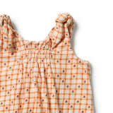 Close-up of a sleeveless baby dress with a smocked bodice and knotted shoulder straps, featuring an orange and cream gingham pattern with small red rose motifs, on a white background.