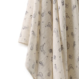 A close-up of a cream-colored muslin fabric, likely a baby blanket or swaddle, featuring a delicate black line-art print of dandelions and bees. The fabric is draped elegantly against a clean white background, highlighting its soft texture and fine pattern details.