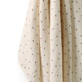 A close-up of a cream-colored muslin fabric, likely a baby blanket or swaddle, featuring a small brown embroidered floral pattern and soft vertical folds.
