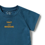 A close-up of a teal blue short-sleeve t-shirt with the French phrase 'TOUT est MAGIQUE' printed in gold on the chest. The inner collar features a small silver graphic of two cartoon faces.