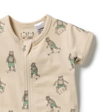 A close-up of a baby romper in a warm cream color, featuring an all-over print of teddy bears wearing green shorts and riding skateboards. The romper has a front zipper and short sleeves, with a visible Camper brand tag inside the collar.