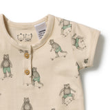 A close-up of a baby short-sleeve henley romper in cream, featuring an all-over print of teddy bears in green shorts skateboarding. The garment has a three-button placket and a visible brand tag reading 'CAUTION KEEP WAY FROM ANY'.