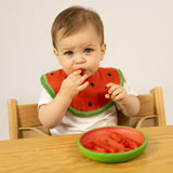 Baby wearing a watermelon-themed bib and eating from a watermelon-shaped plate.
