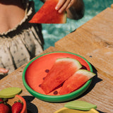 Watermelon slices on a green plate with a person holding a watermelon slice in the background.