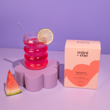 Stacked cups with pink liquid, lime slice, watermelon wedge, and 'mini + me' packaging on a purple background