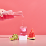 Pink drink being poured into a glass with a watermelon slice, lime, and bottle on a pink background