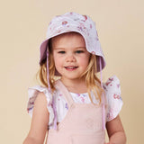 A young girl with blonde hair smiling while wearing a light purple bunny-print sun hat and a pale pink overall dress against a solid beige background.