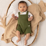 A baby lying on their back in a woven oval bassinet, wearing olive green overall shorts over a white short-sleeve shirt with a colorful floral print. The bassinet is lined with white fabric and draped with a beige checkered muslin blanket.
