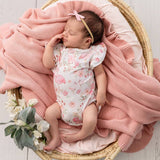 A sleeping newborn baby wearing a pink flamingo print bodysuit and a pink bow headband, resting inside a woven basket lined with a pink knit blanket and cushion.