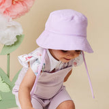 A toddler wearing a light lavender bucket hat with chin straps, a white ruffle-sleeve top with a butterfly print, and matching lavender shorts, posed against a beige background with large decorative paper flowers.