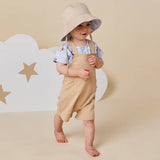 A toddler standing barefoot in a studio setting, wearing a beige bucket hat with a patterned underside, a light blue short-sleeve shirt, and beige overall-style shorts. The background is a warm beige with white cloud shapes and star cutouts.