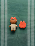 A small doll-shaped charm with a red hat and white outfit stands next to a red apple-shaped accessory on a green striped fabric background.