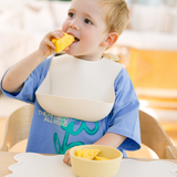 Child wearing a bib eating fruit with a bowl of fruit on a table.