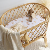 A natural rattan oval bassinet with a cream knit blanket and a white fitted sheet printed with tiny teddy bear motifs. A small stuffed bunny toy sits inside the bassinet on a soft neutral background.