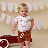 Child wearing a patterned shirt and brown shorts standing in front of a red toy car with a decorative banner in the background.