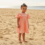 A young girl stands barefoot on a sandy beach wearing a light peach-pink gingham dress with white lace trim at the neckline and hem.