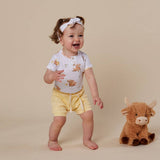 A baby girl wearing a white short-sleeve henley shirt with a brown bear print, yellow shorts, and a white bow headband, standing next to a brown plush toy bull on a beige background.