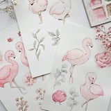 A styled flat-lay of watercolor flamingo and floral art prints on white paper, with pink flamingos, roses, leaves, and small dried flowers arranged around a watercolor palette.