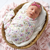 A sleeping newborn baby swaddled in a white muslin swaddle blanket with a colorful wildflower print, resting in a woven rattan basket on a pink blanket. The baby is wearing a matching pink bow headband.