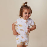 A toddler girl wearing a white short-sleeve zip-front romper with a soft brown animal print, standing against a plain beige studio background.