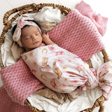 A sleeping newborn baby swaddled in a pink and white floral blanket, wearing a matching pink floral headband, resting inside a woven basket with pink textured blankets.