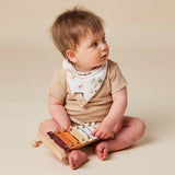 A baby sitting on a beige studio floor, wearing a tan short-sleeve bodysuit and a white patterned bib, while playing with a wooden xylophone toy.