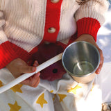 Person holding a metal cup and white spoon with a red and beige sweater, sitting on a star-patterned blanket.