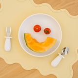 White bowl with a smiley face made of fruit on a yellow placemat with cutlery.