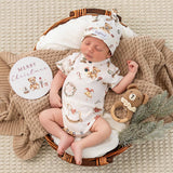 A sleeping newborn baby is posed in a wicker basket on a beige knit blanket, wearing a white Christmas-themed short-sleeve bodysuit and matching hat. The outfit has teddy bear and holiday illustrations.