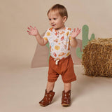A toddler stands in a studio setting wearing a white short-sleeve top with a colorful western-themed print, rust-brown shorts, and brown cowboy boots. The background is beige with a paper cactus and a hay bale prop.