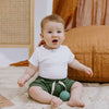 Baby sitting on a carpeted floor with a toy, wearing a white shirt and green shorts.