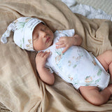 Baby sleeping peacefully on a beige blanket wearing a white hat and white outfit with green patterns.