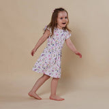 A young girl with a pink hair bow is wearing a white short-sleeve dress with a colorful floral print, posing on a solid beige studio background.