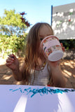 Child holding a colorful cup with a drawing on a table outdoors