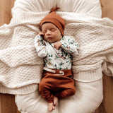 Newborn baby sleeping on a soft surface wearing a floral top and rust-colored pants with a matching headband.