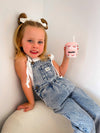 Young girl in denim overalls holding a pink cup with a straw against a plain background